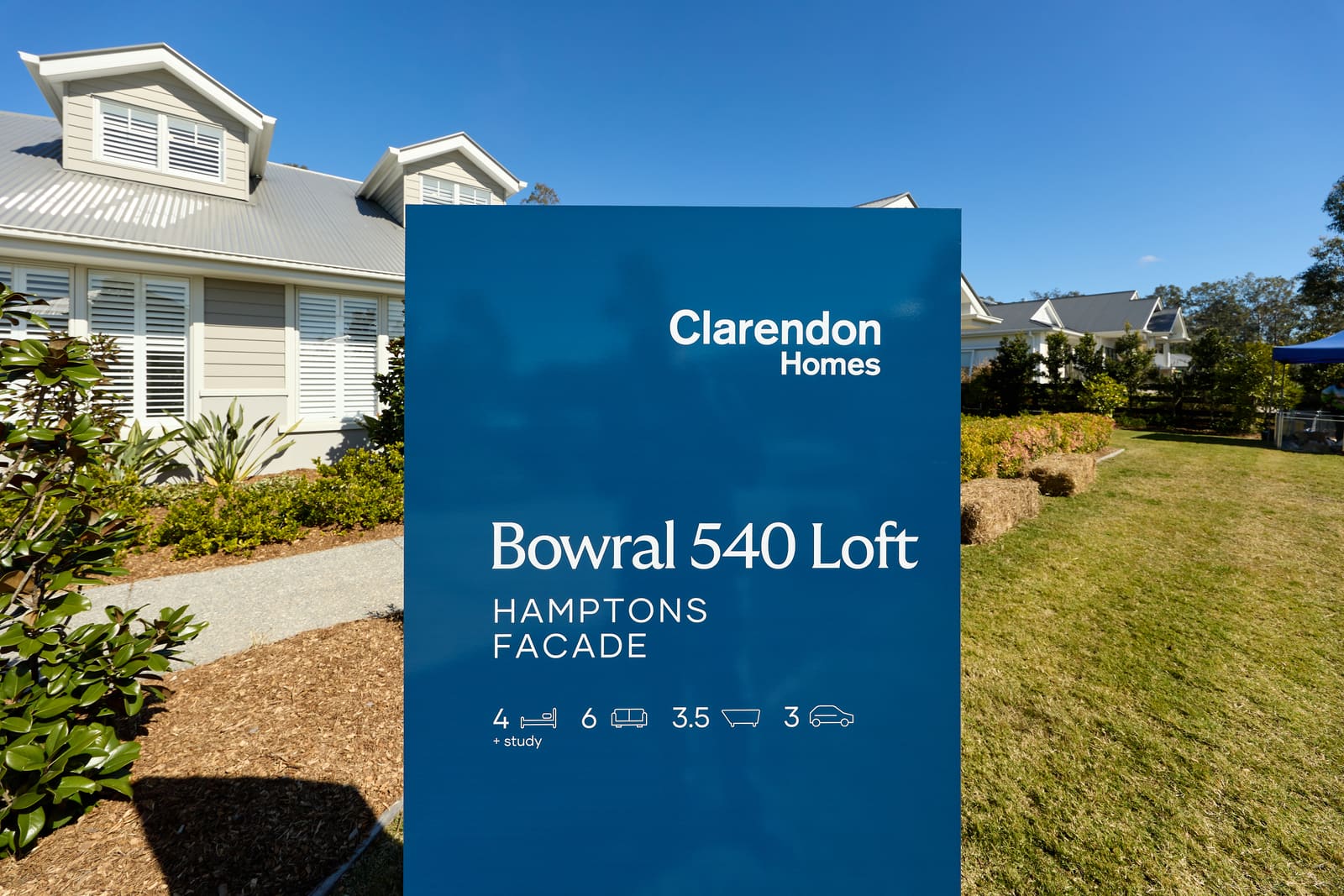 A sign outside a display home at an open day, naming the house design and its bedrooms, bathrooms and garage spaces, with a lawn and more display homes behind it.