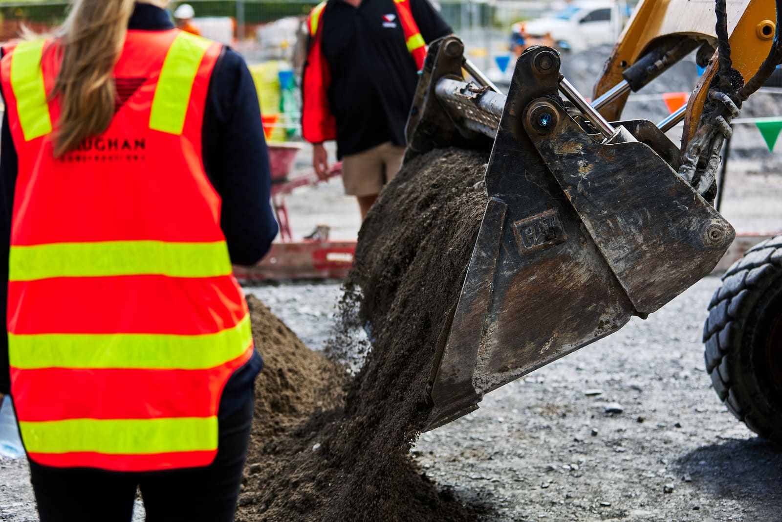 Two people in orange high-visibility vests on a construction site, watching a loader tip a bucket of soil onto a pile.