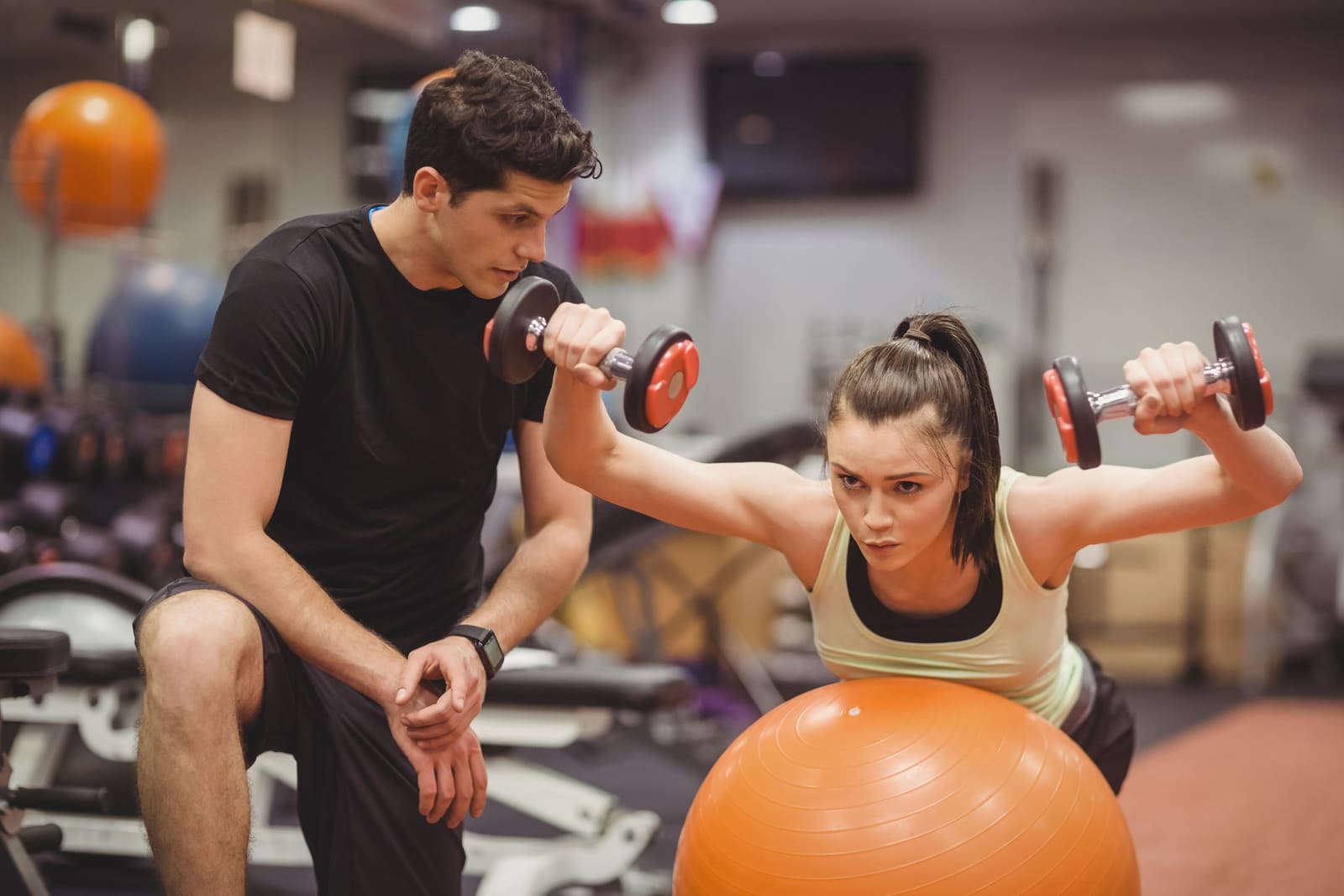 A gym member lifting dumbbells while leaning on an exercise ball, with a trainer kneeling beside them watching their form.