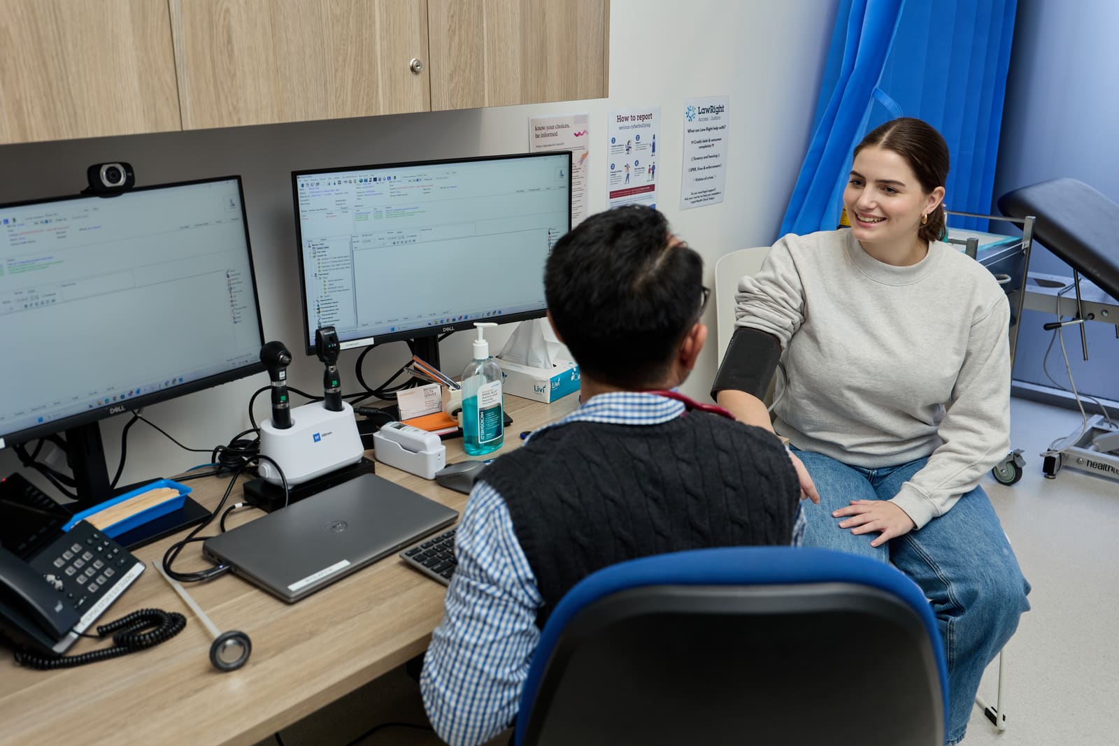 A patient smiling in a consulting room while a doctor takes their blood pressure, beside a desk with two screens.