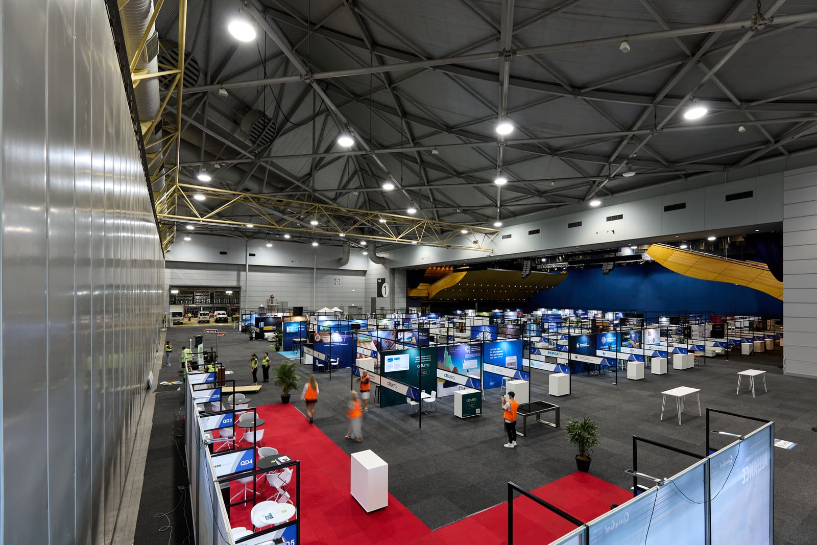 An exhibition hall seen from above while exhibitor stands are being set up, with staff in high-visibility vests walking the aisles.