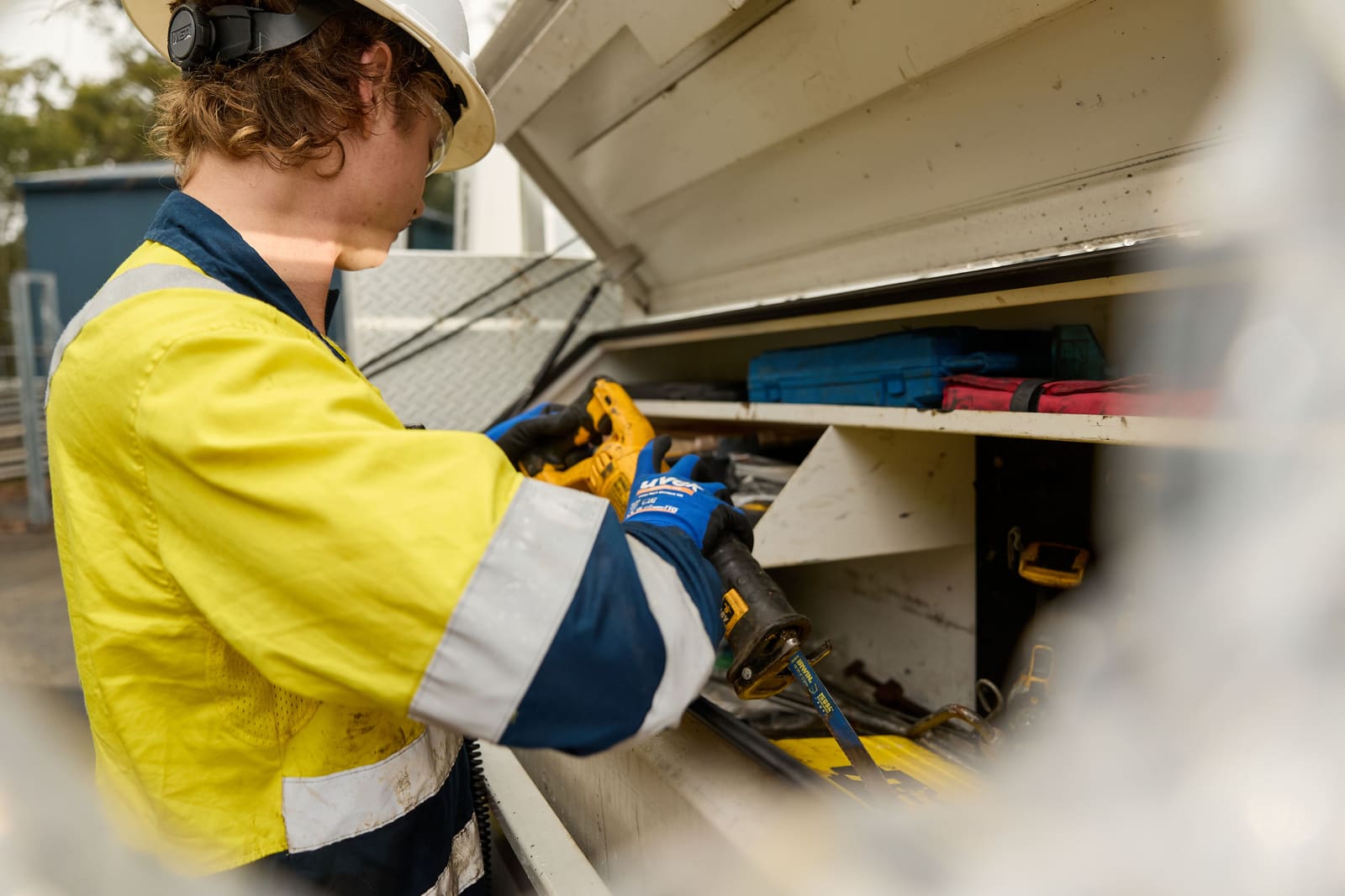 A worker in a hard hat, safety glasses and a high-vis shirt taking a power tool out of the toolbox on the back of a work vehicle.